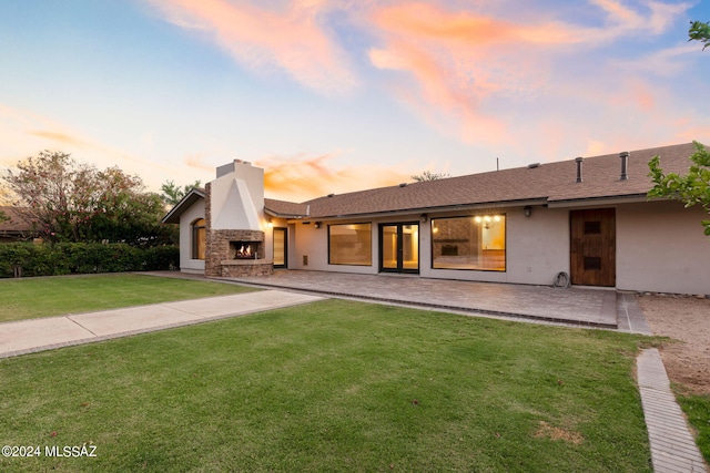 back house at dusk with exterior fireplace, a yard, and a patio