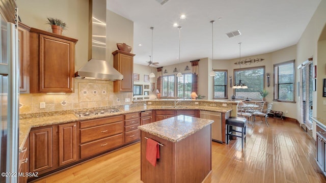 kitchen featuring hanging light fixtures, ceiling fan, wall chimney exhaust hood, a kitchen island, and stainless steel gas cooktop