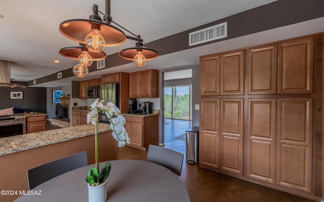 kitchen with appliances with stainless steel finishes, decorative light fixtures, a textured ceiling, wall chimney exhaust hood, and light stone counters