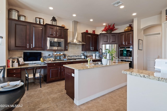 kitchen with wall chimney exhaust hood, stainless steel appliances, backsplash, a center island with sink, and light stone countertops