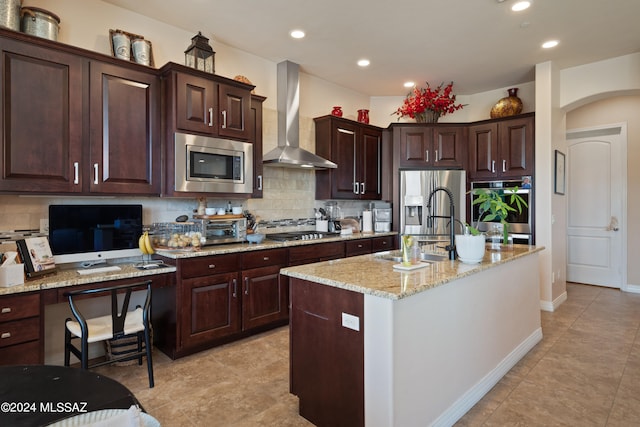 kitchen with light stone countertops, stainless steel appliances, a kitchen island with sink, sink, and wall chimney range hood