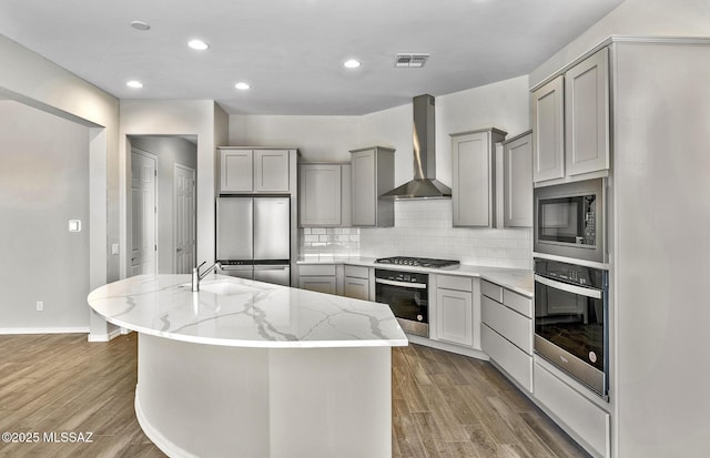 kitchen featuring wall chimney range hood, gray cabinets, appliances with stainless steel finishes, a kitchen island with sink, and light stone counters