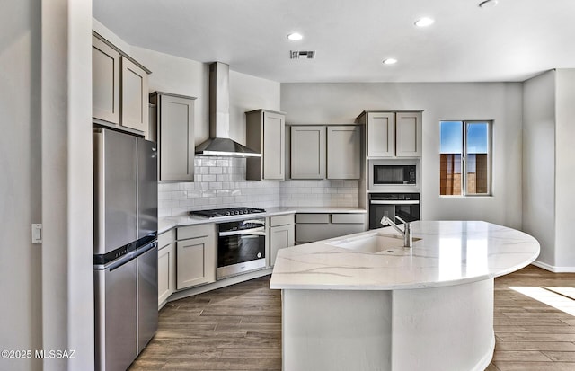 kitchen featuring sink, gray cabinets, stainless steel appliances, an island with sink, and wall chimney exhaust hood