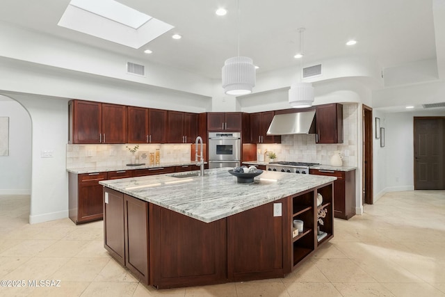 kitchen featuring sink, wall chimney range hood, light stone counters, stainless steel appliances, and a center island with sink