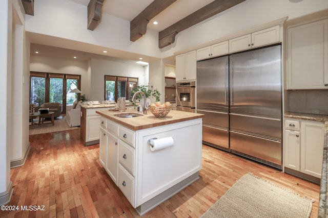 kitchen with wooden counters, a center island with sink, appliances with stainless steel finishes, beam ceiling, and sink