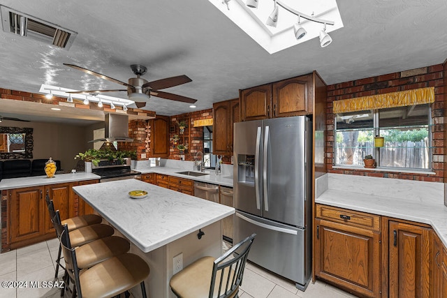 kitchen featuring stainless steel appliances, wall chimney range hood, light tile patterned floors, a kitchen island, and ceiling fan