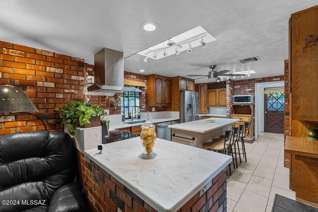 kitchen featuring stainless steel appliances, island range hood, rail lighting, brick wall, and ceiling fan