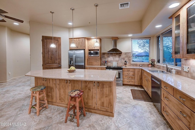 kitchen with wall chimney range hood, appliances with stainless steel finishes, backsplash, a kitchen island, and a kitchen bar