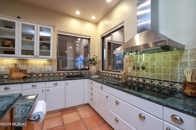 kitchen with wall chimney range hood, white cabinets, backsplash, sink, and black cooktop