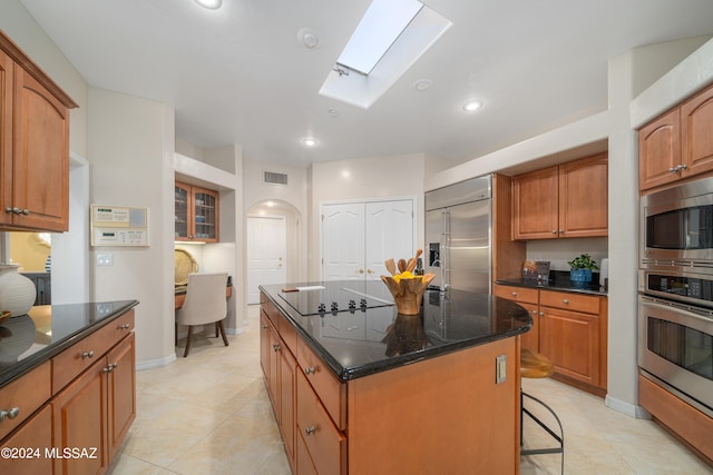 kitchen with a skylight, light tile patterned flooring, a kitchen island, built in appliances, and dark stone countertops