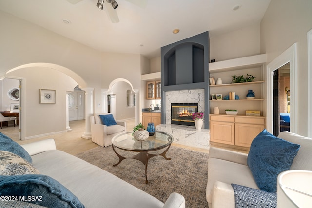 living room with built in features, ceiling fan, ornate columns, a premium fireplace, and high vaulted ceiling