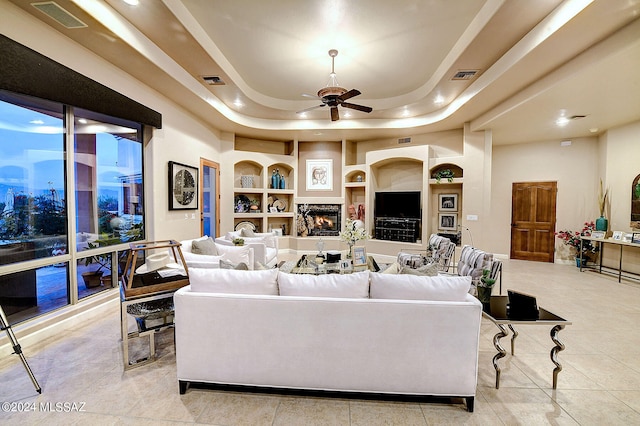 living room with ceiling fan, a raised ceiling, built in features, and light tile patterned floors