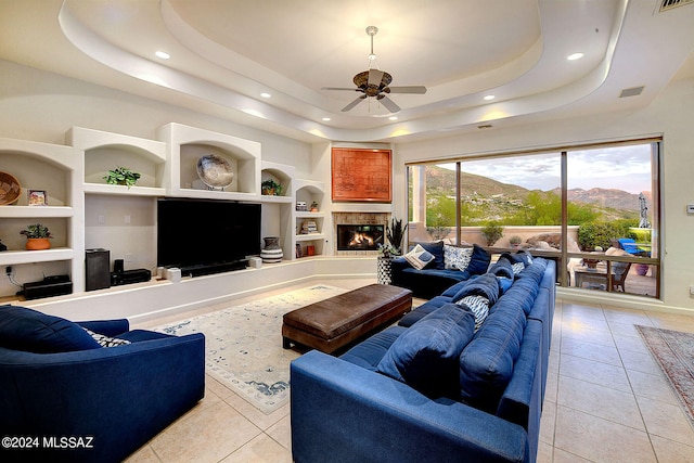 tiled living room with ceiling fan, a raised ceiling, a tile fireplace, and built in features