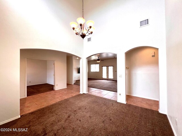 carpeted empty room featuring ceiling fan with notable chandelier, a high ceiling, tile patterned floors, arched walkways, and visible vents