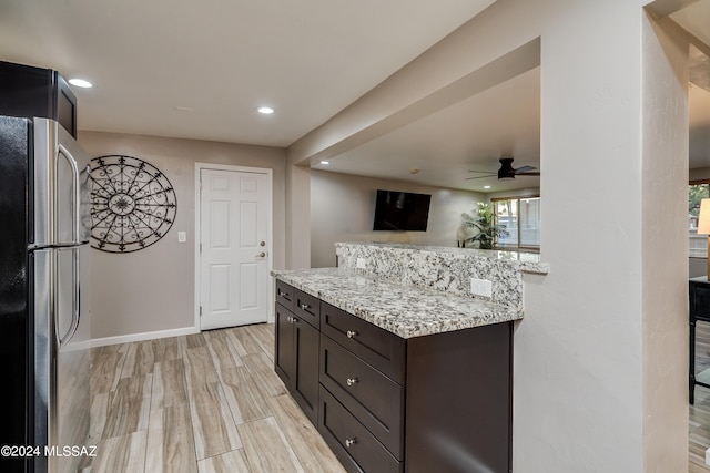 kitchen with dark brown cabinets, light hardwood / wood-style floors, light stone countertops, stainless steel refrigerator, and ceiling fan