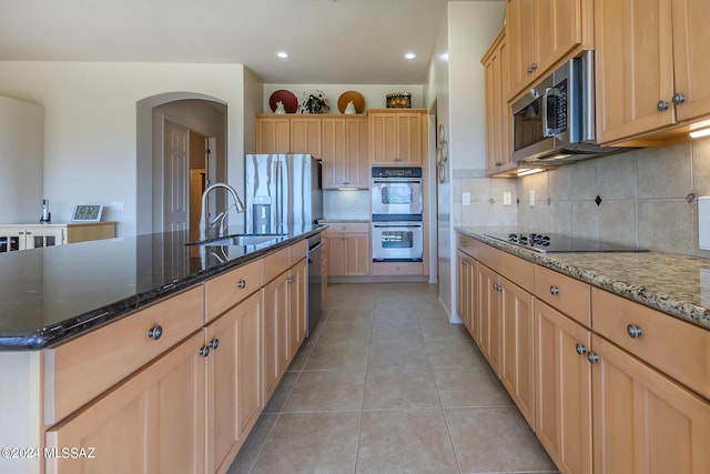 kitchen featuring sink, light tile patterned floors, dark stone countertops, stainless steel appliances, and an island with sink