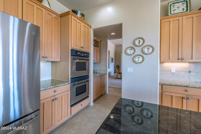 kitchen featuring light tile patterned floors, appliances with stainless steel finishes, backsplash, light brown cabinetry, and dark stone counters