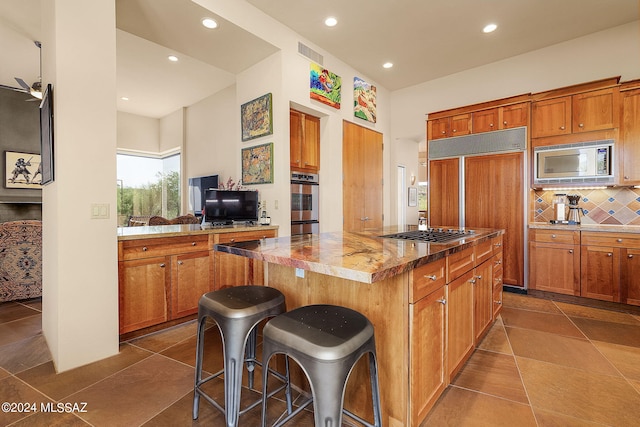 kitchen with backsplash, light stone counters, built in appliances, a kitchen island, and a breakfast bar