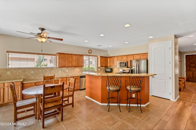 kitchen with stainless steel appliances, a center island, backsplash, a ceiling fan, and light stone counters