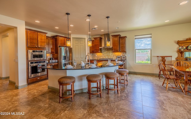 kitchen featuring a center island with sink, decorative backsplash, appliances with stainless steel finishes, dark countertops, and wall chimney exhaust hood