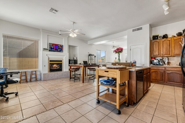 kitchen with ceiling fan with notable chandelier, a tile fireplace, a center island with sink, black dishwasher, and light tile patterned flooring