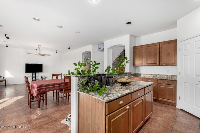 kitchen with light tile patterned flooring, light stone counters, a kitchen island, a ceiling fan, and brown cabinetry