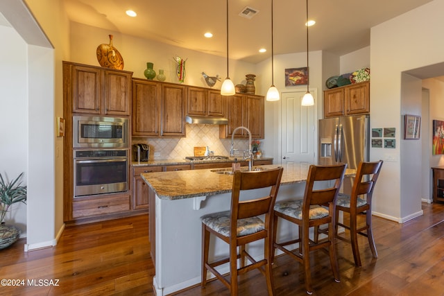 kitchen with light stone countertops, appliances with stainless steel finishes, decorative backsplash, decorative light fixtures, and a center island with sink