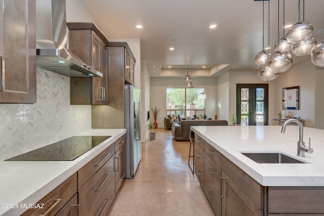 kitchen with a kitchen island with sink, stainless steel fridge, sink, hanging light fixtures, and wall chimney range hood