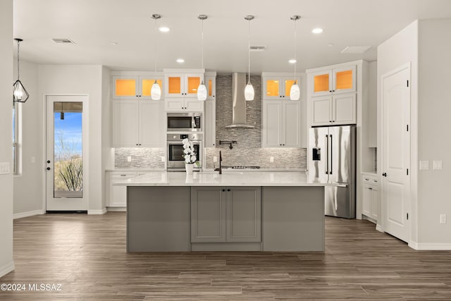 kitchen with appliances with stainless steel finishes, wall chimney exhaust hood, a kitchen island with sink, white cabinetry, and hanging light fixtures