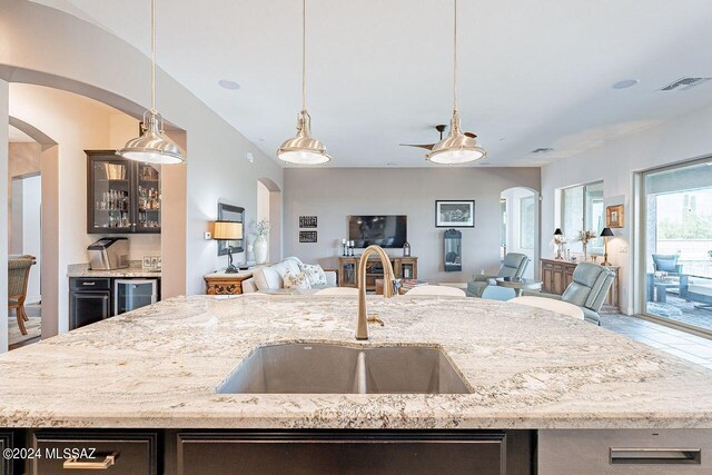 kitchen with decorative light fixtures, beverage cooler, an island with sink, light stone countertops, and sink