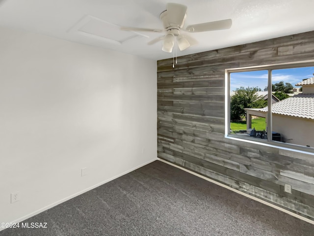 carpeted empty room with ceiling fan, an accent wall, wood walls, baseboards, and attic access