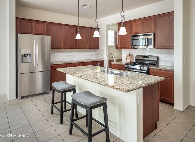 kitchen featuring light stone countertops, a kitchen island with sink, decorative light fixtures, backsplash, and stainless steel appliances