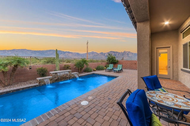 pool at dusk featuring a mountain view, a jacuzzi, a patio area, and pool water feature