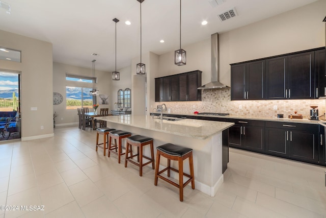 kitchen with sink, decorative light fixtures, a kitchen island with sink, wall chimney range hood, and a breakfast bar area