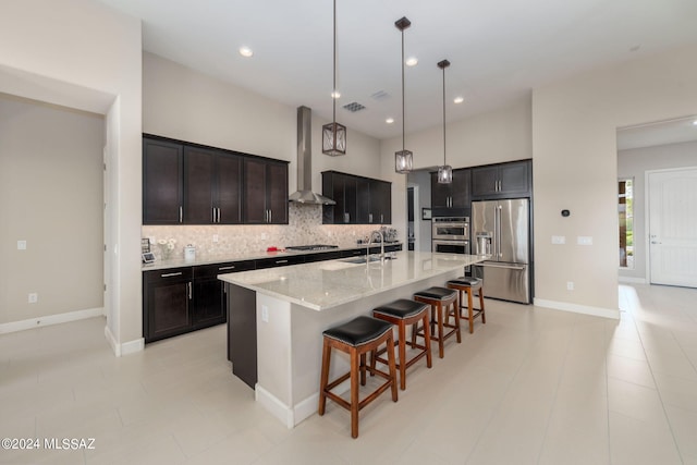 kitchen with appliances with stainless steel finishes, an island with sink, backsplash, wall chimney exhaust hood, and decorative light fixtures