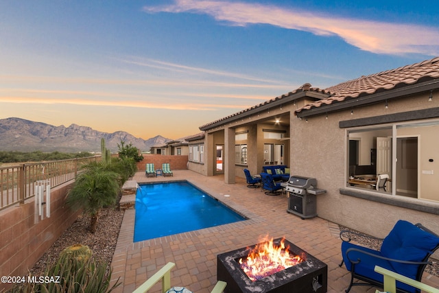 pool at dusk featuring a patio, a mountain view, area for grilling, and an outdoor fire pit