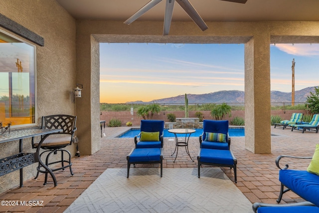 patio terrace at dusk featuring ceiling fan, a mountain view, and a fenced in pool