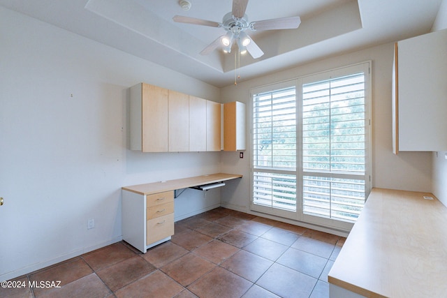 unfurnished office featuring built in desk, a raised ceiling, light tile patterned floors, and ceiling fan