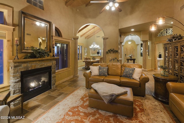 living room with a stone fireplace, beam ceiling, decorative columns, a high ceiling, and ceiling fan with notable chandelier