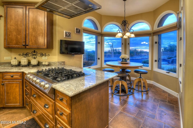 kitchen with brown cabinets, light stone counters, stainless steel gas stovetop, an inviting chandelier, and extractor fan