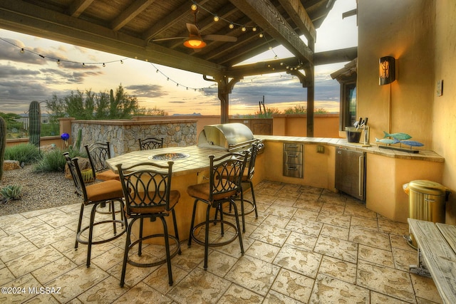 patio terrace at dusk with ceiling fan, outdoor wet bar, area for grilling, and a grill