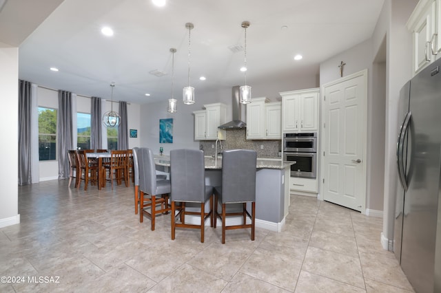 kitchen with hanging light fixtures, an island with sink, stainless steel appliances, light stone countertops, and wall chimney range hood