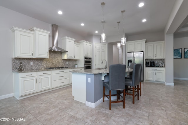 kitchen with sink, a kitchen island with sink, light stone counters, stainless steel appliances, and wall chimney exhaust hood