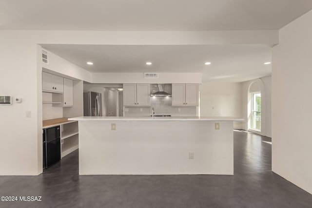 kitchen featuring decorative backsplash, wall chimney exhaust hood, an island with sink, stainless steel fridge, and gray cabinets