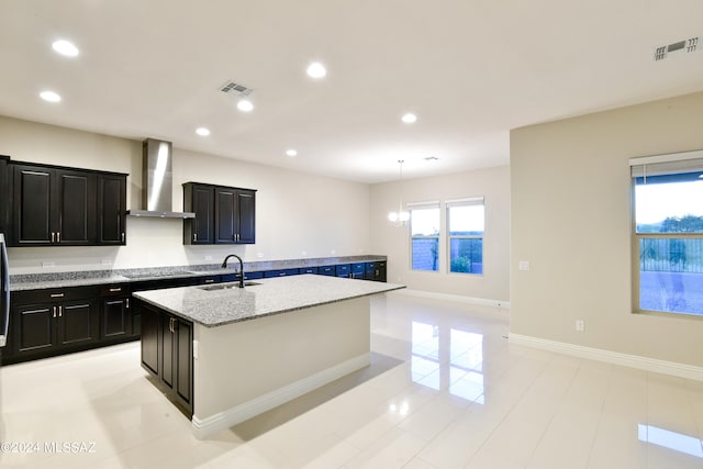 kitchen with visible vents, dark cabinets, a kitchen island with sink, wall chimney range hood, and a sink