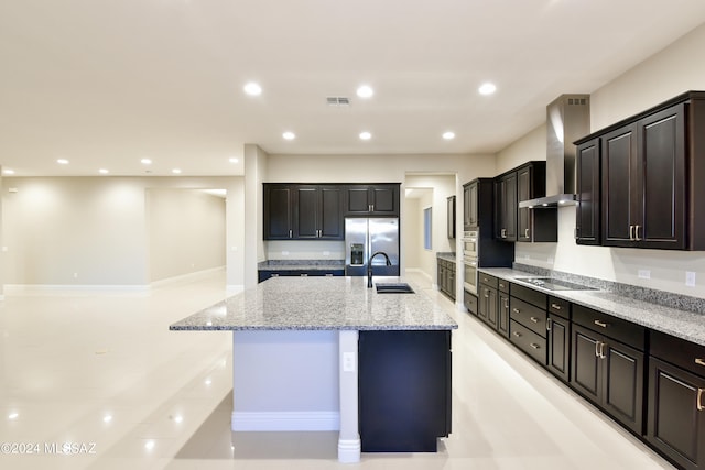 kitchen featuring light stone counters, wall chimney range hood, appliances with stainless steel finishes, dark brown cabinetry, and a kitchen island with sink