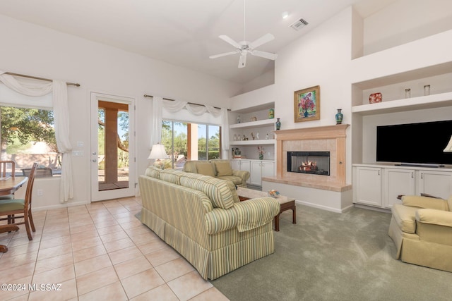 living room featuring light carpet, high vaulted ceiling, built in shelves, a tiled fireplace, and ceiling fan