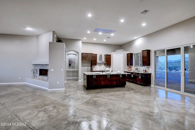 kitchen with dark brown cabinets, wall chimney exhaust hood, a kitchen island with sink, and sink