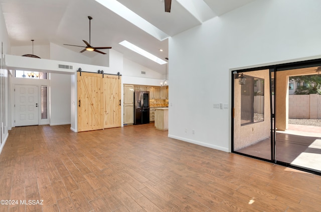 unfurnished living room featuring a barn door, high vaulted ceiling, light hardwood / wood-style flooring, a skylight, and ceiling fan