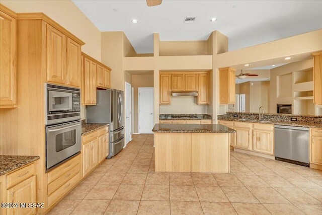 kitchen with ceiling fan, stainless steel appliances, a center island, light brown cabinetry, and dark stone counters